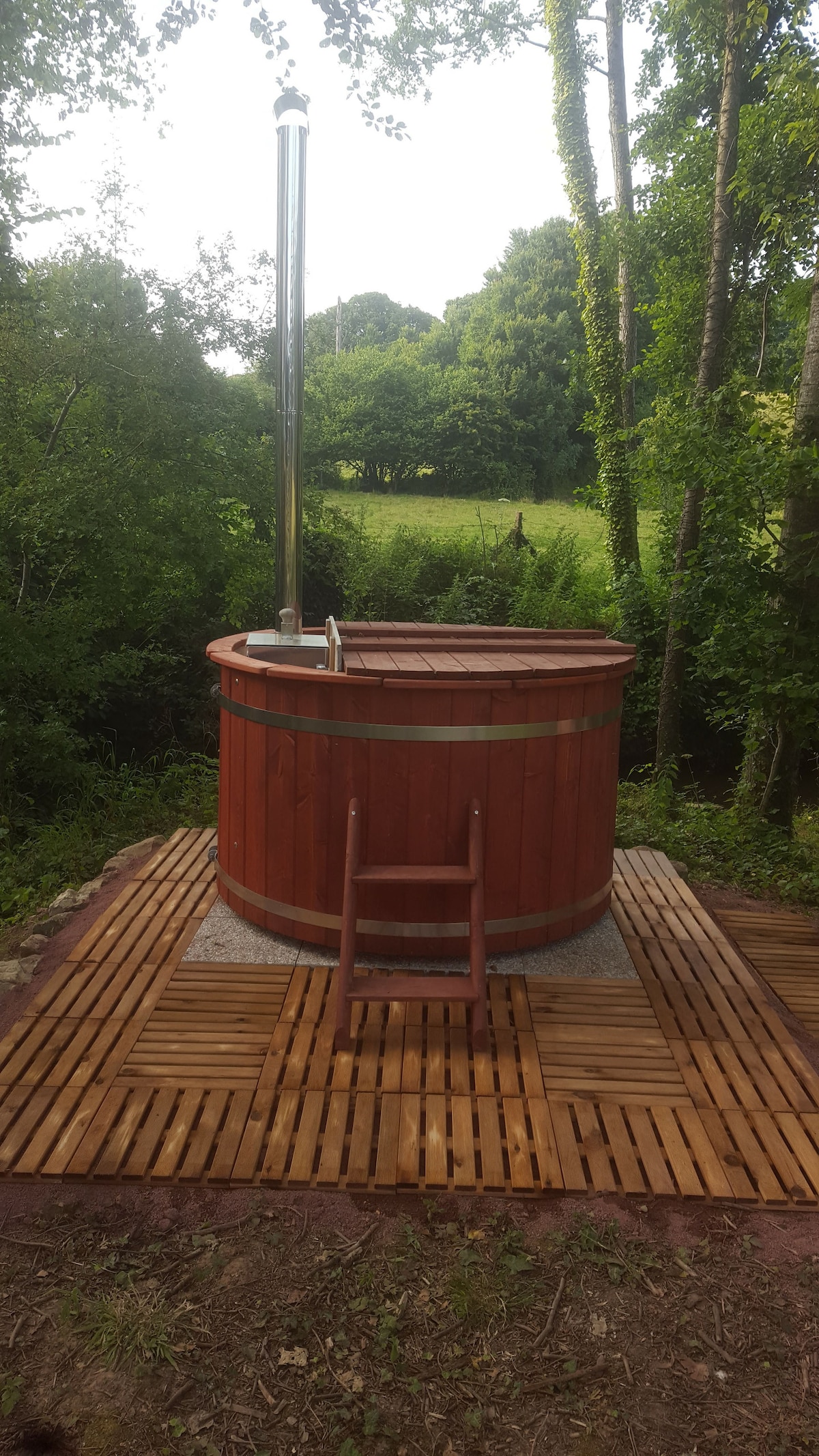 A wooden Nordic bath is positioned on a raised wooden deck, surrounded by lush greenery. A metal chimney extends from the bath, and a wooden ladder provides access. The scene features a serene backdrop of trees and open fields.