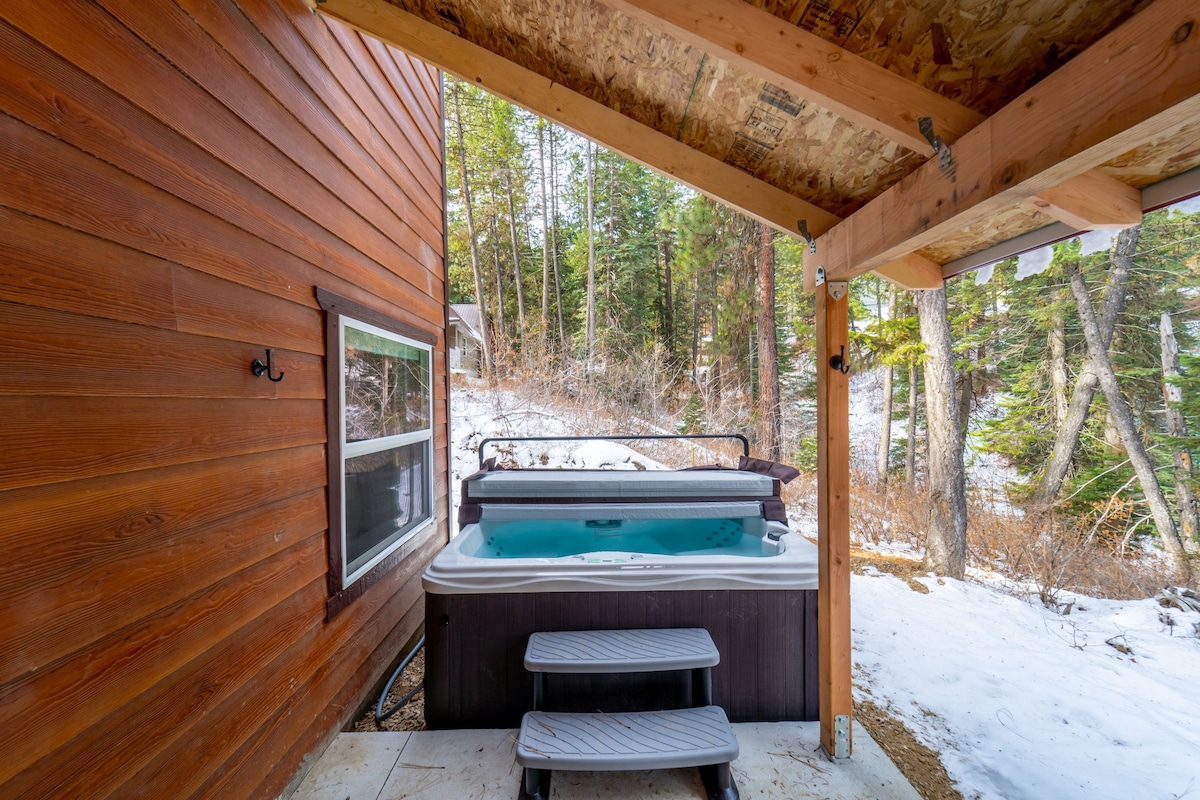 A hot tub is situated on a covered patio, surrounded by a snowy landscape and dense trees. Steps lead up to the hot tub, offering a view of the serene outdoor surroundings. The wooden exterior of the cabin is visible in the backdrop.