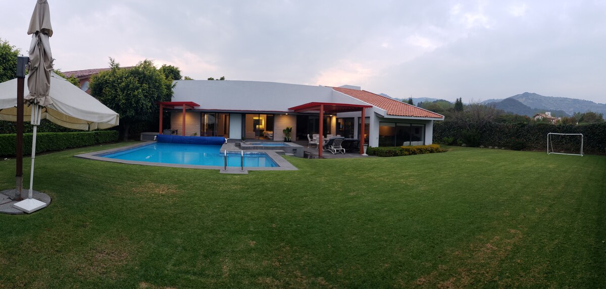 A spacious backyard is presented, showcasing a well-maintained lawn bordered by lush greenery. A private pool is visible, accompanied by patio furniture and a shaded area under an umbrella. The single-story house features large windows reflecting the surrounding landscape.