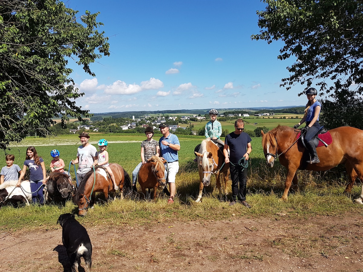 A group of individuals stands in a grassy area, with ponies and horses calmly positioned nearby. The background features rolling hills and a clear blue sky. Several participants are wearing helmets, contributing to a safe riding experience. A dog is seen in the foreground.