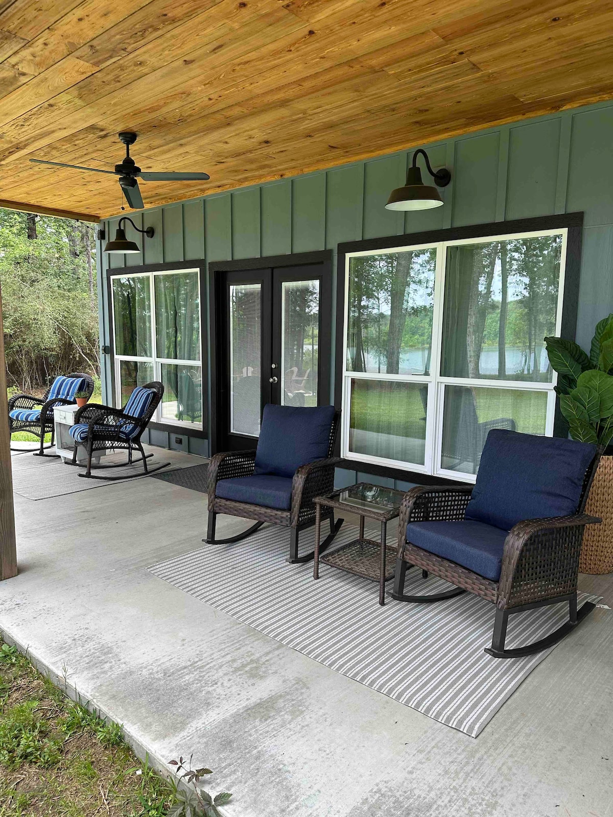 A covered porch features two comfortable chairs with dark cushions facing large windows. A small glass table sits between the chairs on a striped rug. A ceiling fan is mounted above, and greenery from the surrounding area is partially visible.