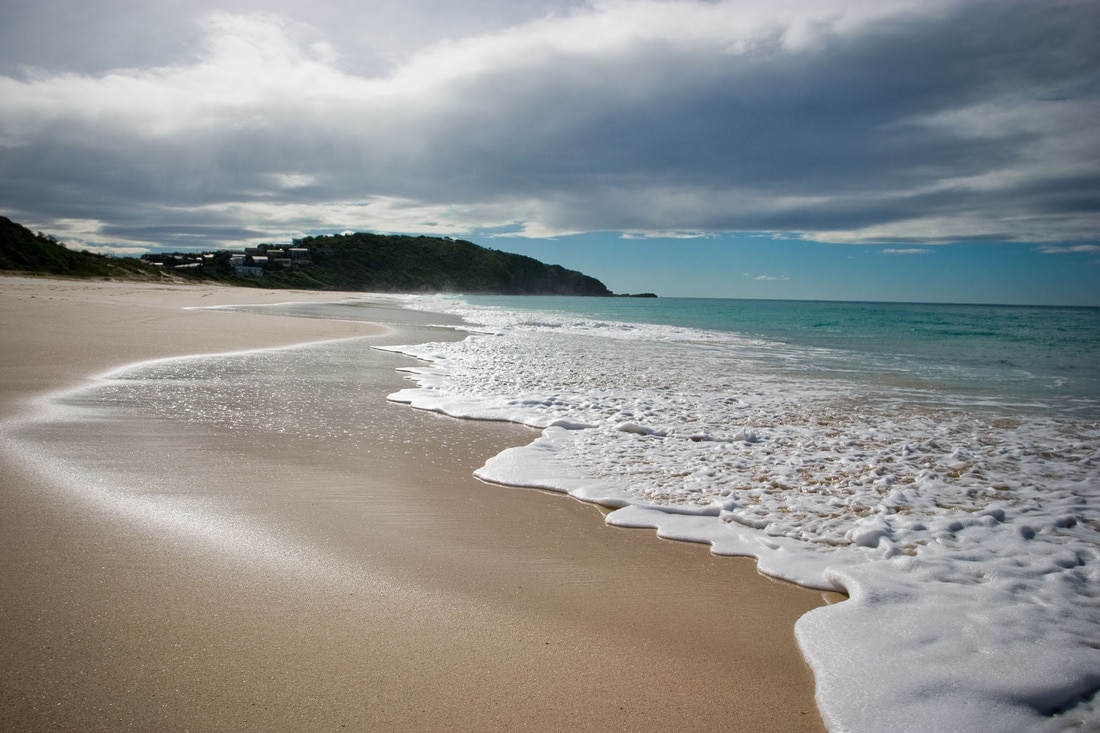 A serene beach scene captures gentle waves lapping at a sandy shore. The coastline curves into the distance, revealing lush greenery and a few seaside homes on a hillside. The sky is partly cloudy and bright, reflecting a tranquil atmosphere by the ocean.
