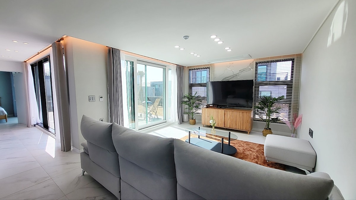 A contemporary living area is presented with large glass doors that allow natural light. A comfortable gray sofa faces a stylish television unit, complemented by indoor plants. Light-colored walls and elegant flooring enhance the spacious feel of the room.