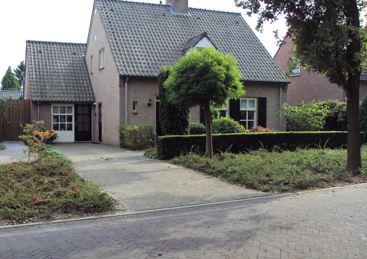 A two-story brick house is surrounded by well-maintained shrubs and trees. A gravel path leads to the entrance, which features a single door and a large window. Lush greenery enhances the inviting outdoor space, complementing the home’s simple yet charming architecture.