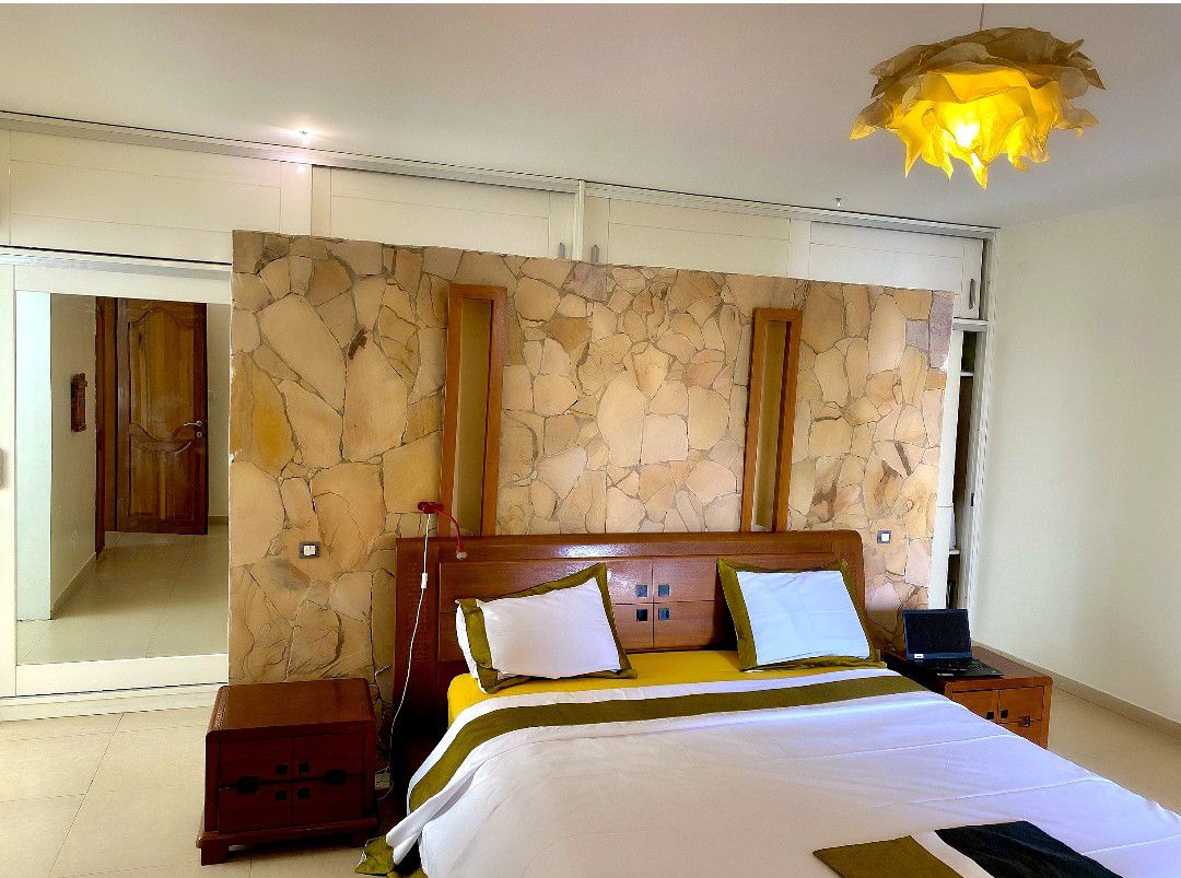 A spacious bedroom is characterized by a stone accent wall, complemented by wooden nightstands on either side of the bed. Soft lighting emerges from a unique ceiling fixture. The bed features crisp white linens and a decorative throw. Large mirrors enhance the sense of space.