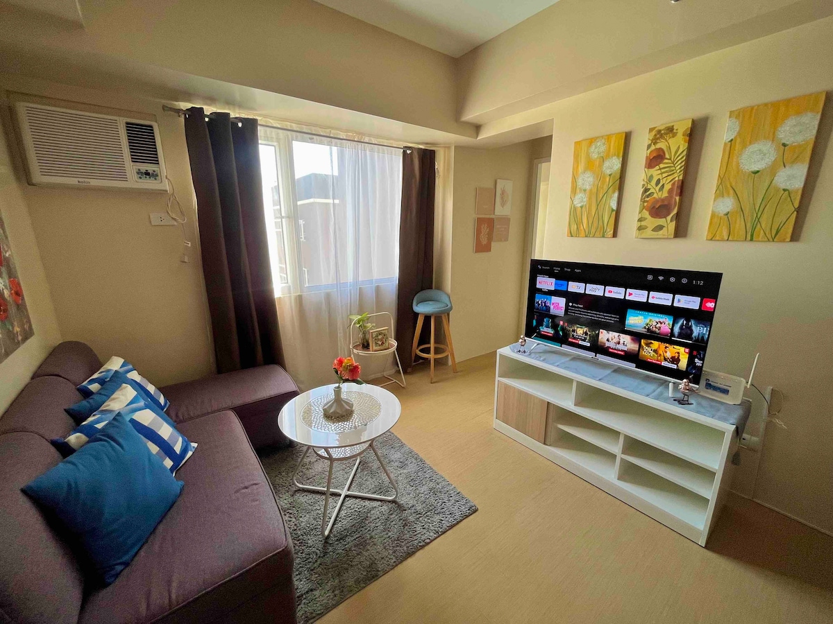 A welcoming living area features a plush sofa adorned with blue cushions and a round coffee table. A television stands on a white console, showcasing various entertainment options. Natural light filters through the window, which is dressed in dark curtains, enhancing the space's comfort.