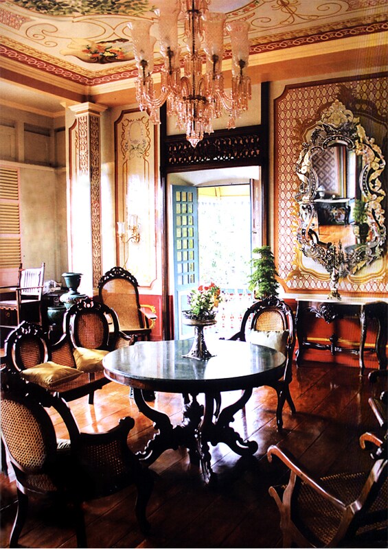 An elegant dining area features a round marble table surrounded by ornate wooden chairs. A decorative chandelier hangs from the ceiling, and a large, intricately framed mirror reflects the room's charming details. Natural light enters through a set of double doors, complemented by potted plants.