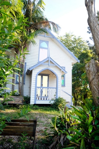 The exterior of a charming white building with blue accents is framed by lush greenery. A large arched entrance leads to a small balcony with decorative railings, nestled among the tropical plants and trees. Natural light enhances the inviting appearance of the structure.