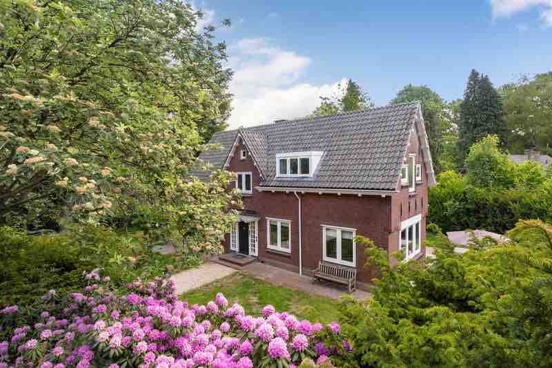 A spacious red-brick house is framed by lush greenery and blooming flowers in the foreground. The structure features multiple windows and a welcoming front porch, creating an inviting exterior. A pathway leads to the entrance, surrounded by a vibrant garden landscape.