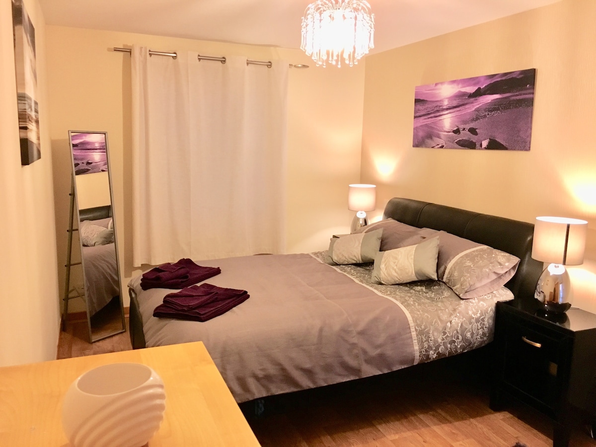 A spacious bedroom is shown, featuring a large bed with gray linens and decorative pillows. Beside the bed, two bedside tables with lamps provide soft lighting. A full-length mirror reflects the room, which is adorned with minimalistic wall art and a window fitted with a light curtain.