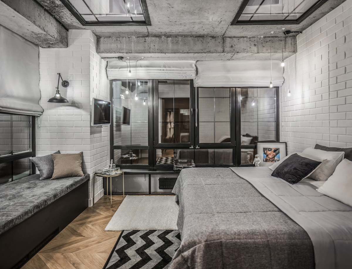 A modern room is displayed with concrete walls and large windows, allowing natural light to fill the space. A spacious bed is layered with gray and white bedding. A cozy seating area is situated by the window, and decorative cushions enhance the ambiance.