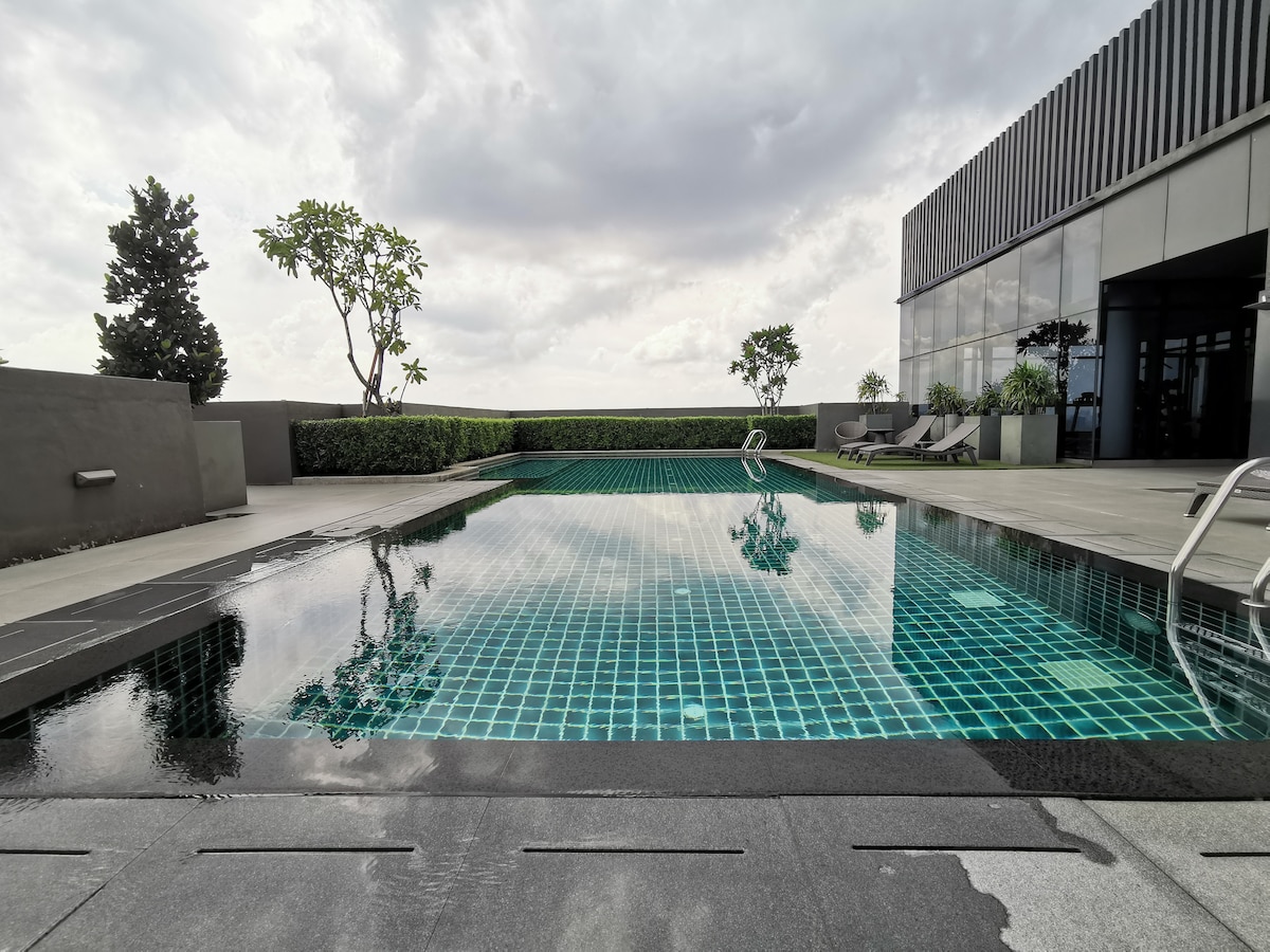 The image features a serene sky pool surrounded by neatly arranged greenery. A sleek tiled surface reflects the sky above, while modern lounge chairs are positioned along the poolside. The area is illuminated by natural light, creating a calm and inviting atmosphere.