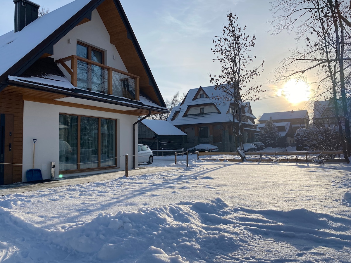 The exterior of a wooden chalet is seen blanketed in fresh snow, with large windows reflecting the soft sunlight. Surrounding houses are visible, and the landscape is punctuated by leafless trees. The serene atmosphere suggests a peaceful winter retreat.