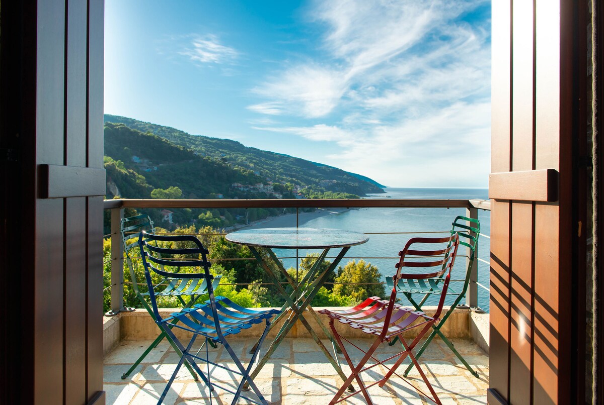 A balcony offers a scenic view of the coastline, framed by open wooden doors. A round table is set with two colorful chairs, one in blue and the other in red, inviting relaxation while overlooking the Aegean Sea and surrounding hills.