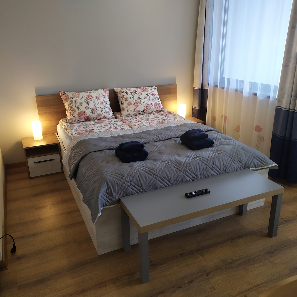 A spacious bed with a light wooden headboard is adorned with floral-patterned bedding. Two bedside lamps offer soft illumination. A small table is placed at the foot of the bed, complemented by a coordinated remote control on its surface. Natural light filters in through a sheer curtain.