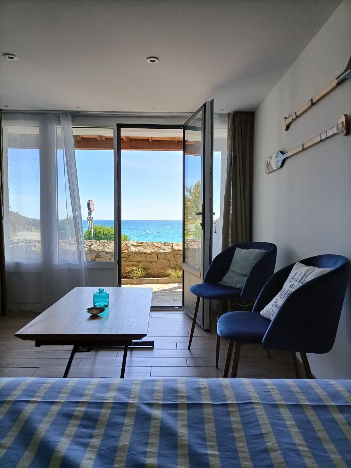 A room is visible with large glass doors opening to a view of the sea. Two blue chairs are placed beside a wooden coffee table. Light curtains allow natural light to fill the space, enhancing the connection with the ocean scenery.