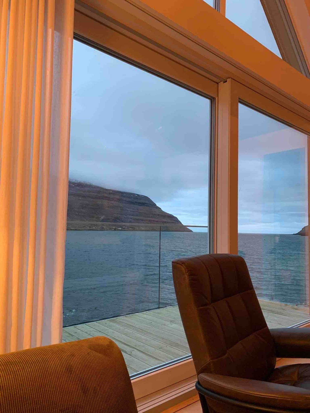 A large window offers a view of the tranquil seafront, framed by light curtains. The distant coastline is visible, with cliffs rising gently from the water. A comfortable chair is positioned nearby, emphasizing the serene atmosphere of the space.
