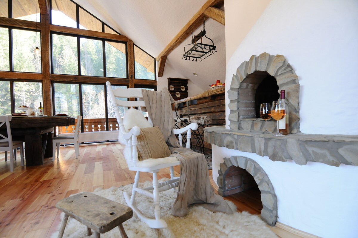 A cozy seating area is highlighted by a white rocking chair and a small wooden stool, positioned near a stone fireplace. Natural light floods the space through large windows, casting a warm glow across the wooden beams and the plush area rug.