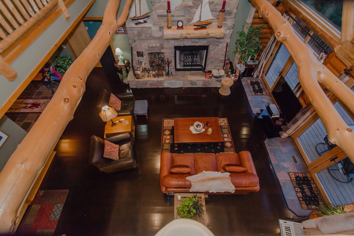 An aerial view of the great room highlights the cathedral ceiling with timber beams. A large fieldstone fireplace is centered against the wall, surrounded by comfortable seating and rustic decor. Warm lighting enhances the cozy atmosphere, while views of the outdoors can be seen through expansive windows.