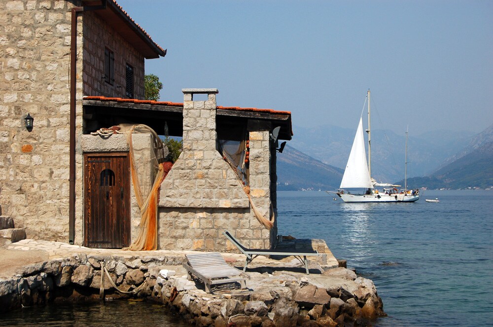 A rustic stone building stands along the water's edge, showcasing the craftsmanship of the past. A sailboat glides gracefully across the calm sea, with distant mountains framing the horizon. Nearby, fishing nets and a sun lounger rest on a stone jetty, inviting relaxation.