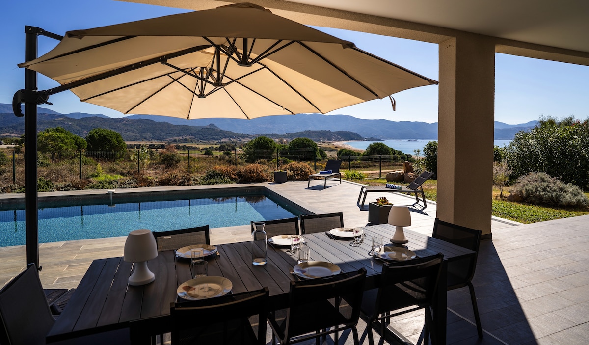 An outdoor dining area is highlighted under a large umbrella, featuring a table set for six. The space offers views of a private pool and the surrounding landscape, with mountains in the background and the sea visible on the horizon.