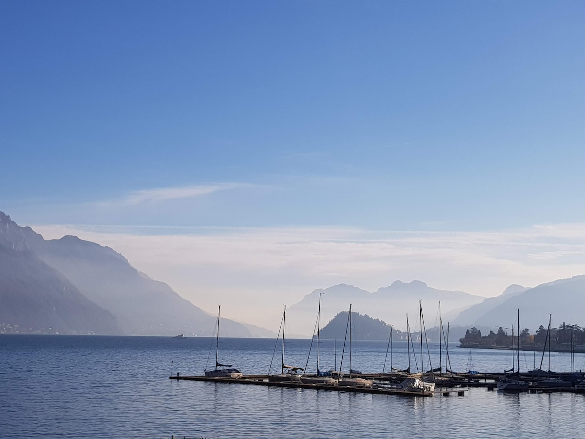 A tranquil view of Lake Como is captured, showcasing a marina with several sailboats moored at the docks. Gentle hills and mountains line the horizon, while a clear blue sky reflects on the water's surface, enhancing the serene atmosphere.