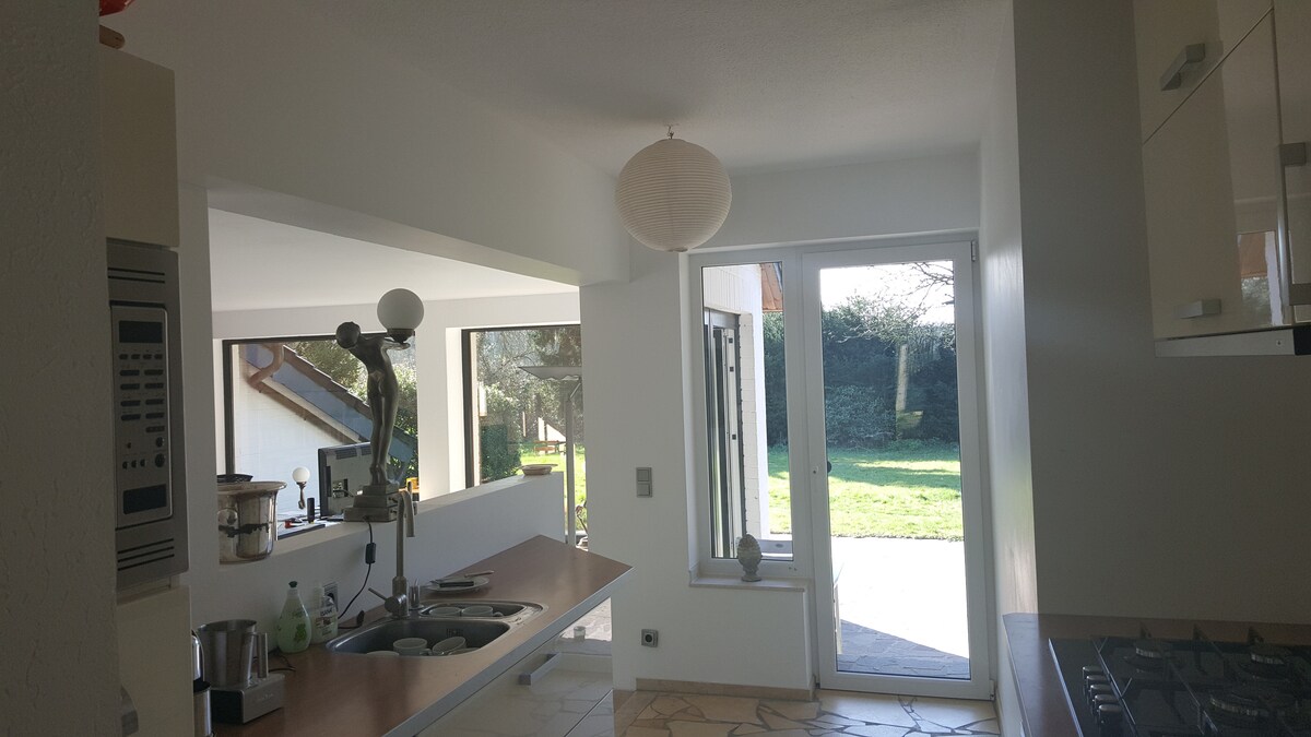 A modern kitchen is visible, featuring a spacious countertop and built-in appliances. Natural light streams in through large windows and a sliding glass door, leading to an outdoor area with greenery. A round paper lantern provides ambient lighting from the ceiling.