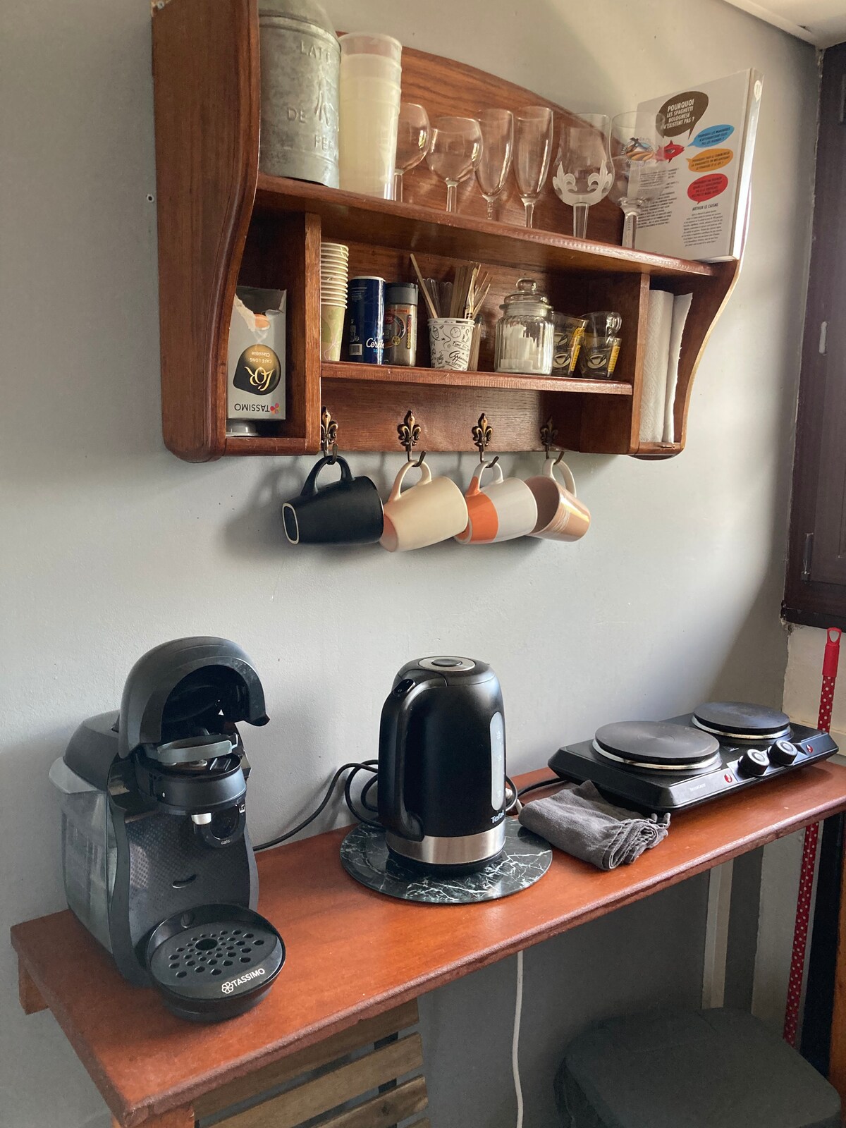 A compact kitchen space features a wooden shelf displaying cups, glasses, and various kitchen items. Below, a coffee maker, a kettle, and a hot plate are arranged on a wooden countertop. The wall is painted in neutral tones, offering a functional and tidy appearance.