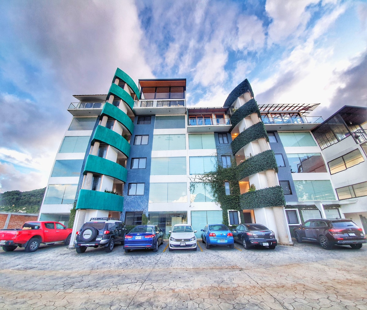 A modern three-story building features a combination of green and blue accents on its facade. Large windows are framed by sleek lines, and a variety of cars are parked in front, indicating accessibility. The rooftop terrace is visible, providing potential outdoor space.