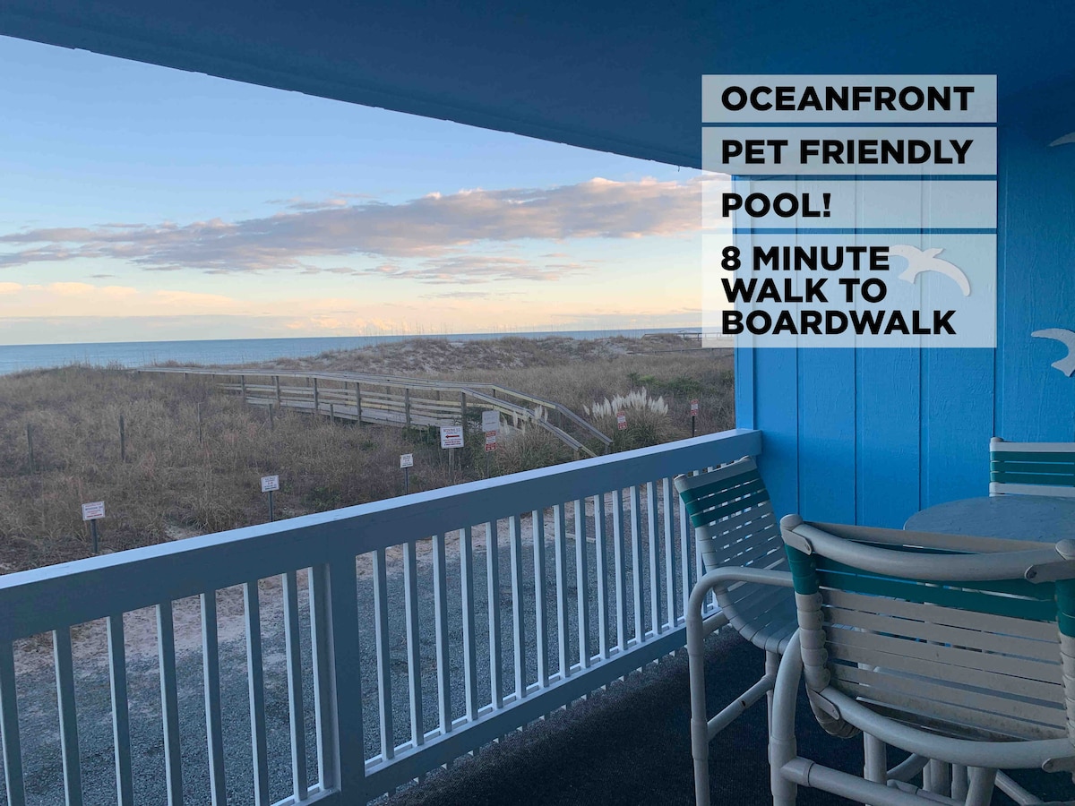 A covered patio area features a round table with four chairs offering a view of the beach and ocean. The railing is painted white, contrasting with the blue walls. An inviting sandy path leads to the beach, surrounded by tall grasses.
