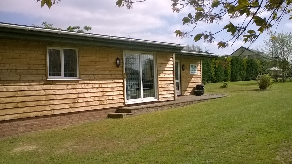 The exterior of the self-catering cabin is displayed, featuring wooden cladding and large glass doors that open to a garden area. Neatly trimmed grass surrounds the cabin, with trees and shrubs in the background. A welcoming, open space is created outside.
