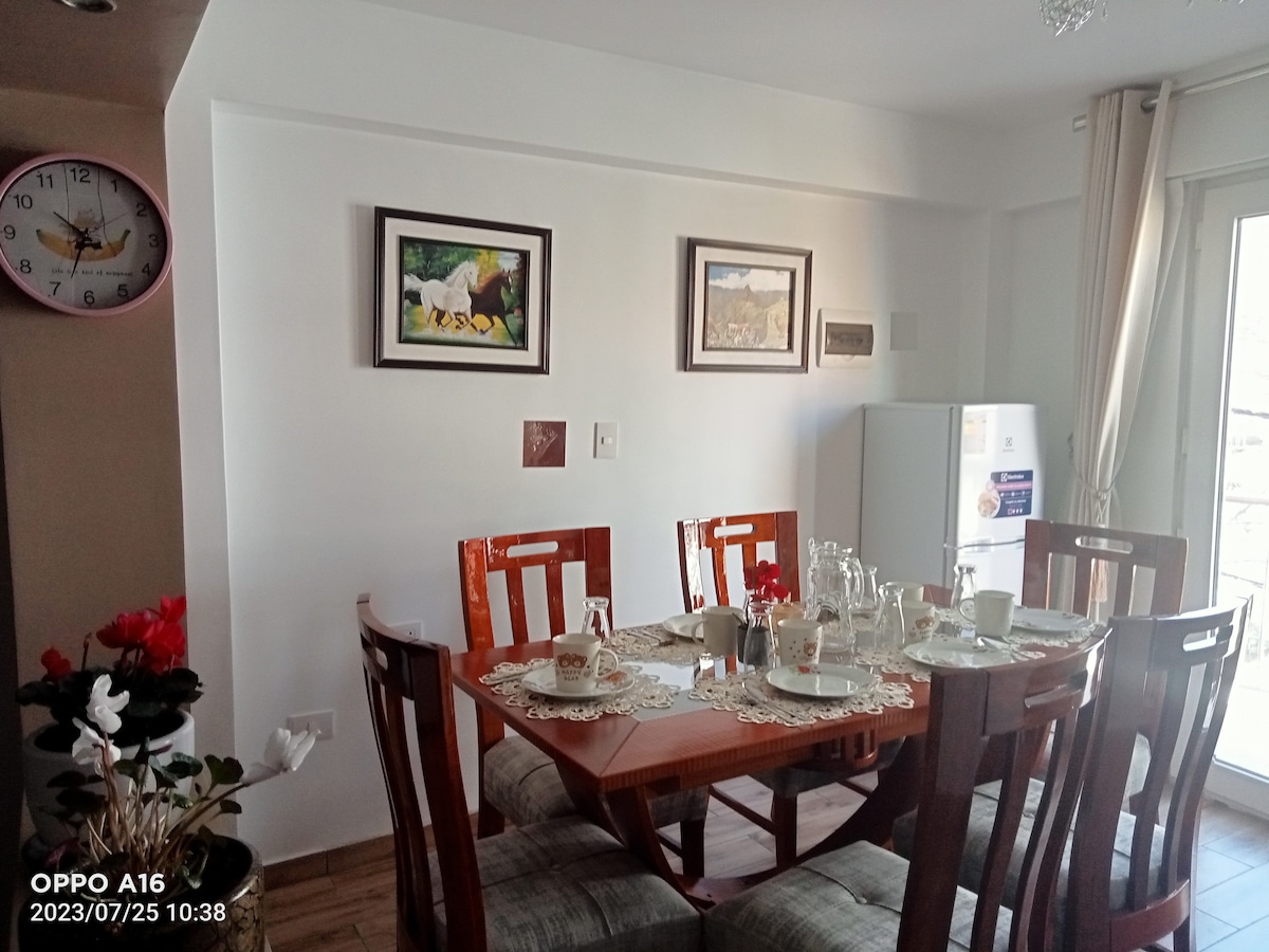 A dining area is presented with a wooden table surrounded by six chairs. The table is set with plates and utensils. Two framed artworks adorn the wall, and a refrigerator is visible to the side, with a window allowing natural light to brighten the space.