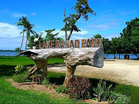 A wooden sign reading 'Lake Danao Park' is prominently displayed, surrounded by a well-maintained landscape featuring lush green grass, tropical plants, and palm trees. The serene lake is visible in the background, under a clear blue sky with scattered clouds.