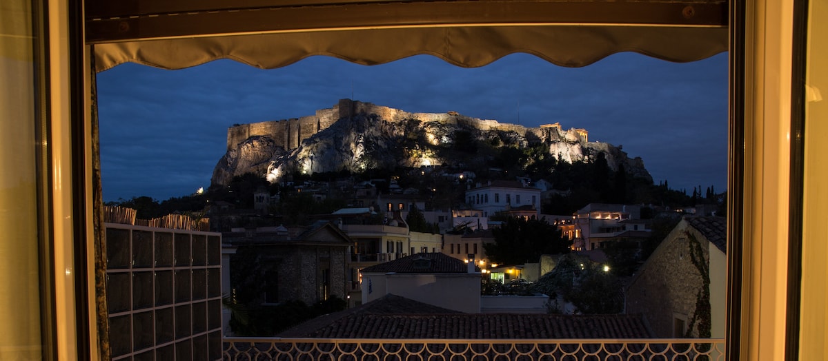The view from the window showcases the illuminated Acropolis against a twilight sky. The soft glow highlights the ancient structure, and the surrounding neighborhood features terracotta roofs and lit streets, creating a serene evening atmosphere.