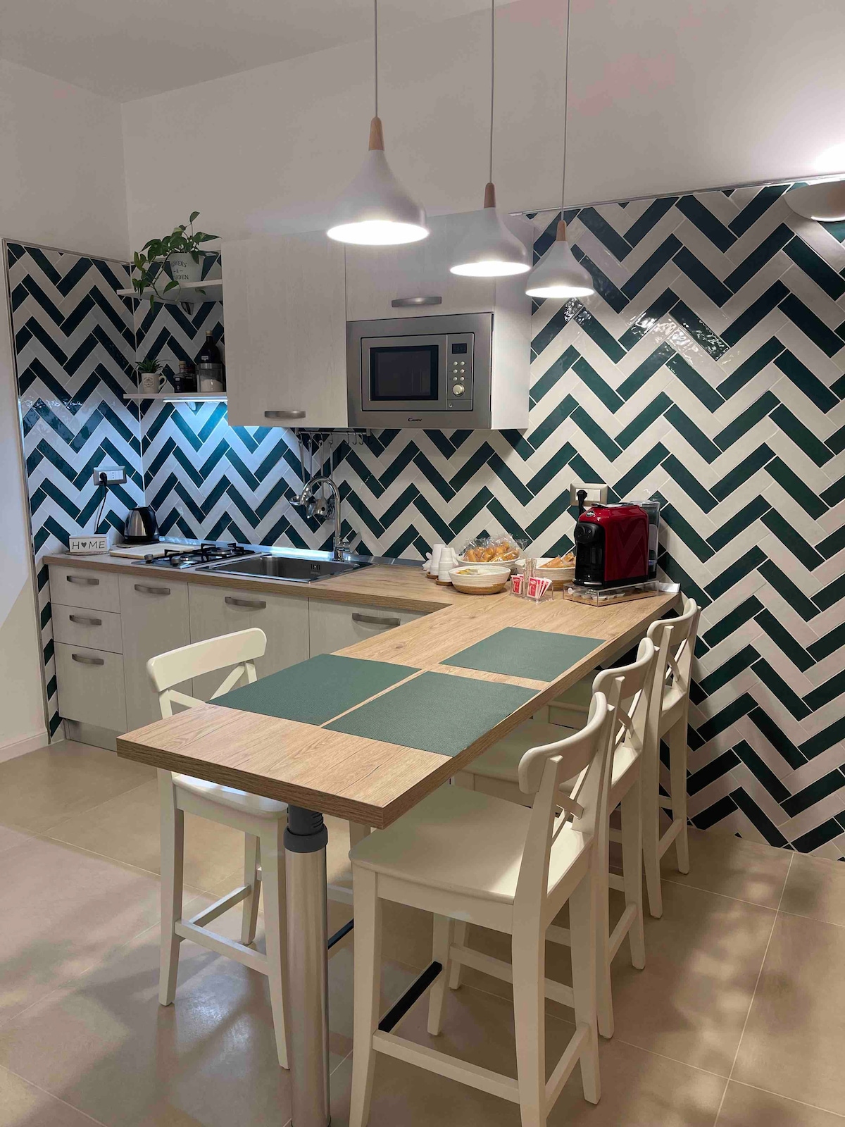 A modern kitchen features a stylish chevron-patterned backsplash in green and white. A breakfast bar with four white stools provides seating. Appliances include a microwave and coffee maker, along with neatly arranged utensils and decorative items, contributing to a functional yet inviting cooking space.