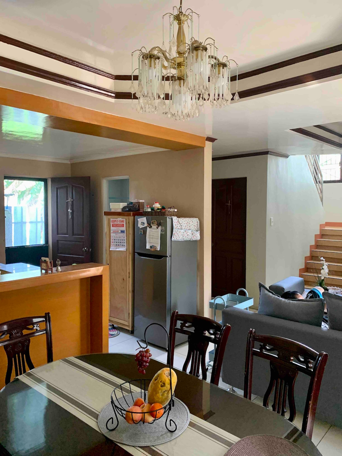 A dining area is featured with a round table surrounded by wooden chairs. A fruit bowl sits on the table, and a chandelier hangs from the ceiling. The kitchen area is visible, with a refrigerator and a wooden cabinet, while a staircase leads to the upper level.
