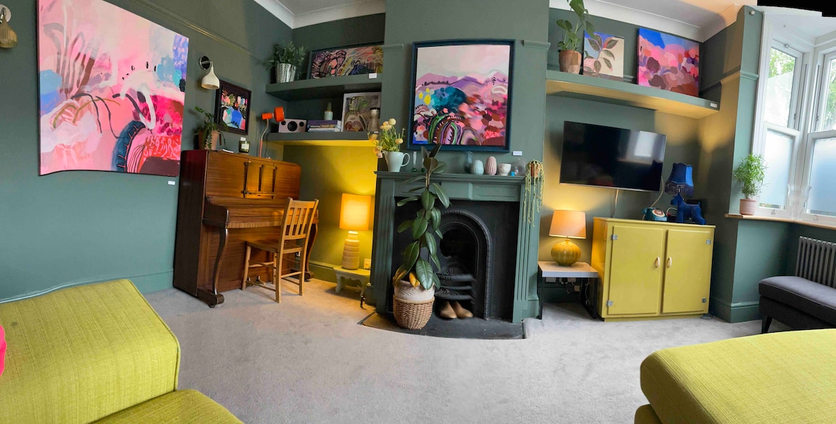The lounge area features a piano and a small wooden chair alongside a vibrant selection of artwork on the walls. Shelves hold decorative items and plants. A television is positioned above a cozy fireplace, complemented by soft lighting from various lamps throughout the room.