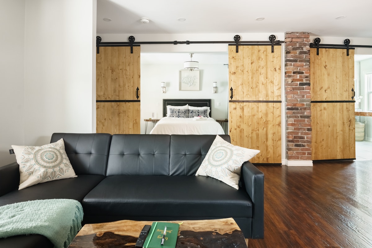 A modern living area features a sleek black sofa adorned with decorative pillows. Sliding cedar barn doors are positioned behind the sofa, leading to a bedroom area. A live edge wood coffee table rests on the polished hardwood floor, enhancing the room’s natural elements.