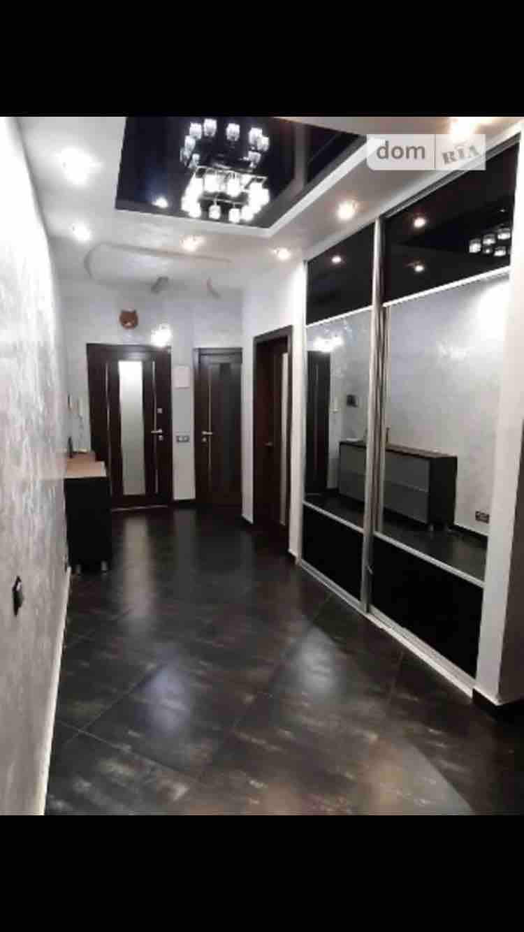 A spacious hallway is featured with a modern design, showcasing a glossy black tile floor. Wall mirrors reflect light and create a sense of openness. The ceiling is adorned with a decorative chandelier, enhancing the elegance of the space. Several wooden doors lead to other areas.