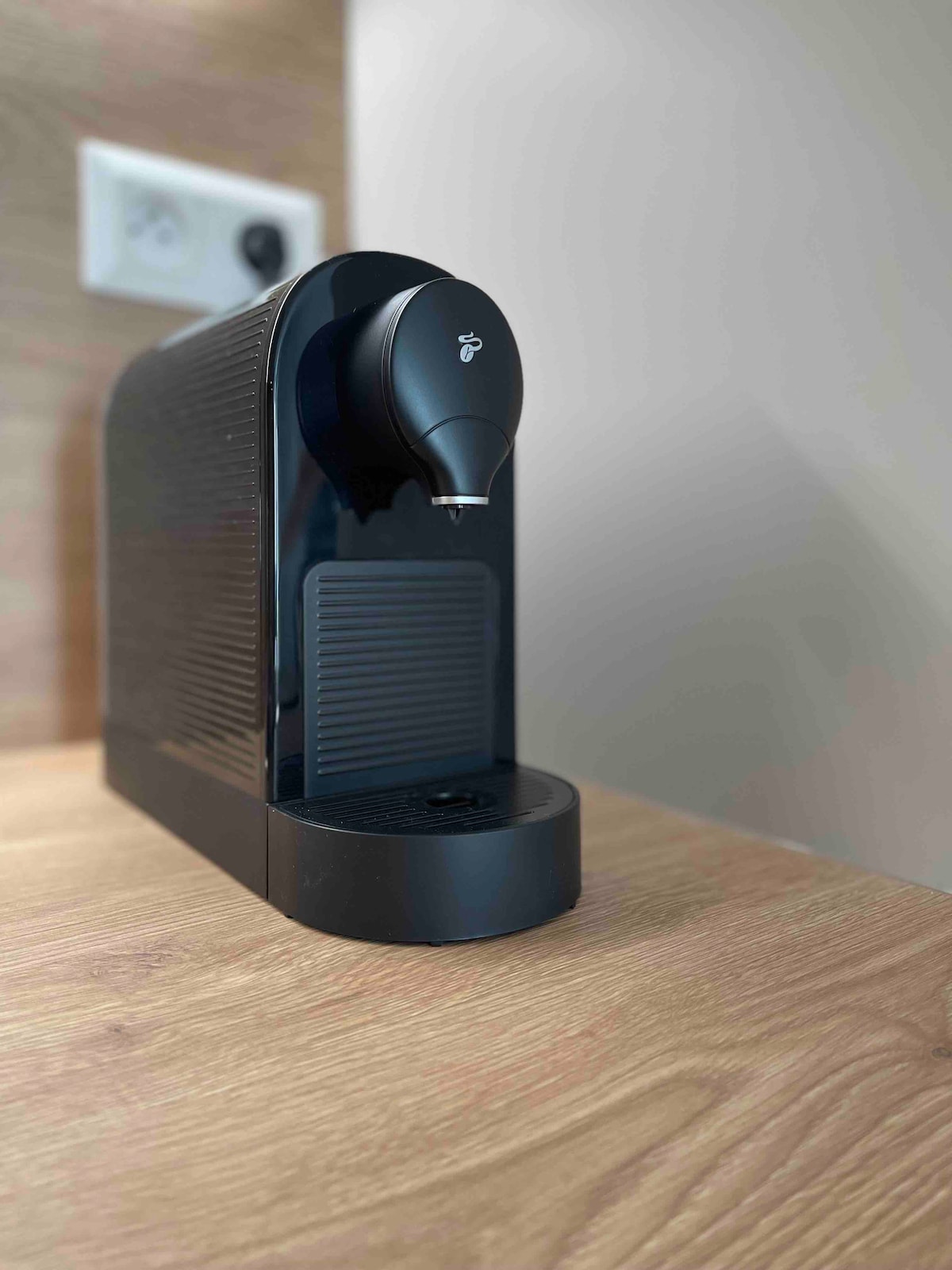 A sleek coffee machine is positioned on a wooden countertop. Its modern design features a prominent black exterior with a smooth finish and subtle branding. A power outlet is visible in the background, indicating ready access for use.