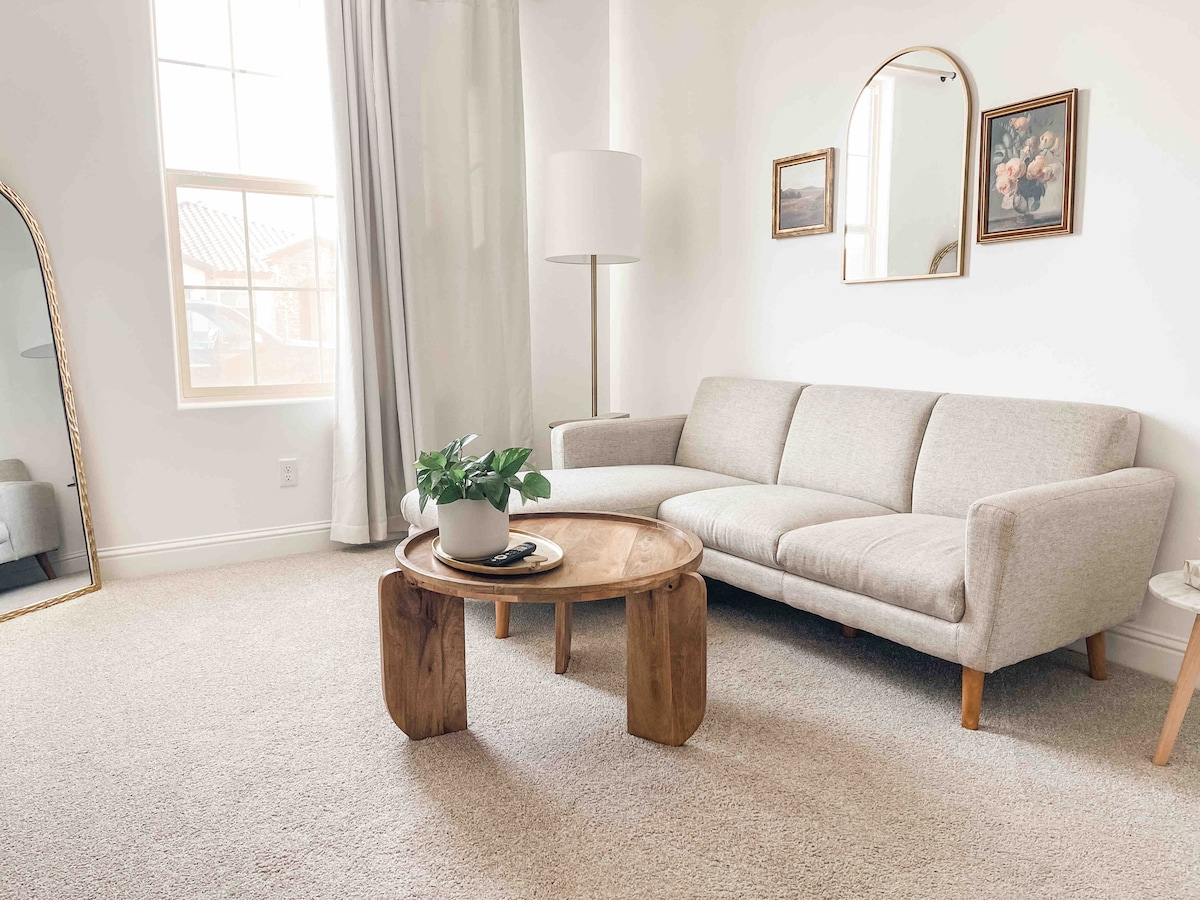 A light-filled living area features a soft gray sofa positioned beside a round wooden coffee table. A potted plant sits atop the table, while a tall lamp provides additional lighting. A large mirror reflects the nearby window, contributing to the space's airy feel.