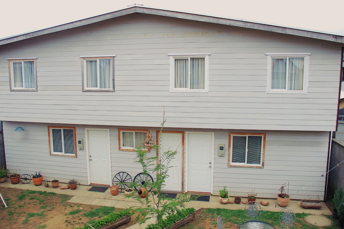 The exterior of a two-story cabaña is displayed, featuring a light-colored wooden facade and multiple windows. Two separate entries are visible, with potted plants adorning the ground. A small garden area is present, adding a touch of greenery to the setting.