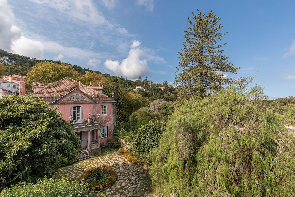 A historic building with a pink façade is surrounded by lush greenery and flowering plants. A stone pathway leads to the entrance, framed by various shrubs and trees. The mountainous backdrop is visible under a clear blue sky, contributing to the serene garden setting.