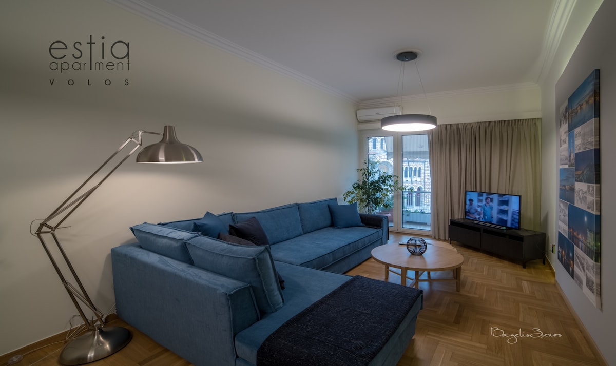 A spacious living room features a large blue couch facing a coffee table and a TV. Sunlight enters through the balcony door, illuminating the area. An indoor plant adds a touch of greenery, while artwork lines the wall, complementing the modern decor.