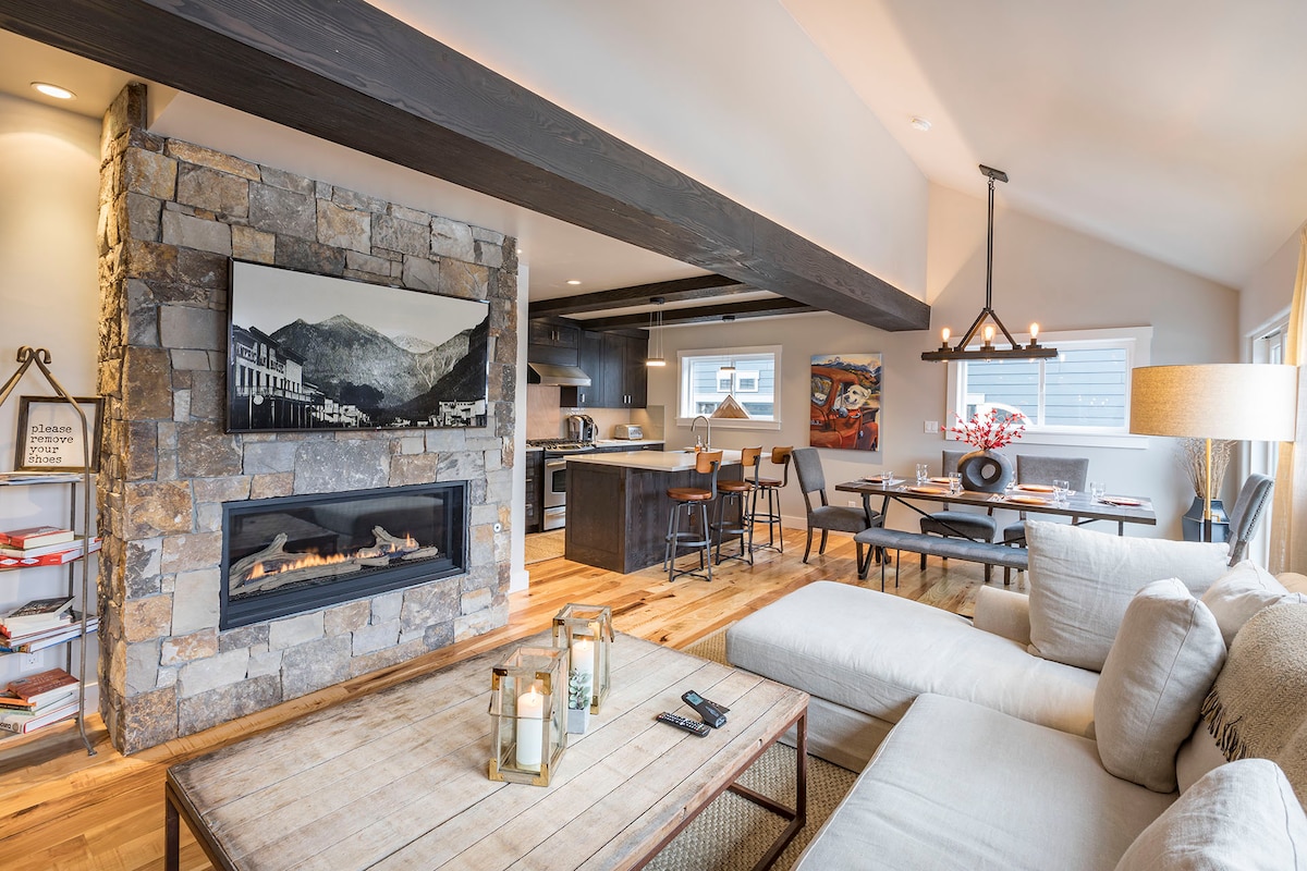 A spacious living area features a stone fireplace with a modern gas insert, surrounded by natural light streaming through large windows. A comfortable sectional sofa complements a wood coffee table, while a dining area is visible with a full-size table and chairs.