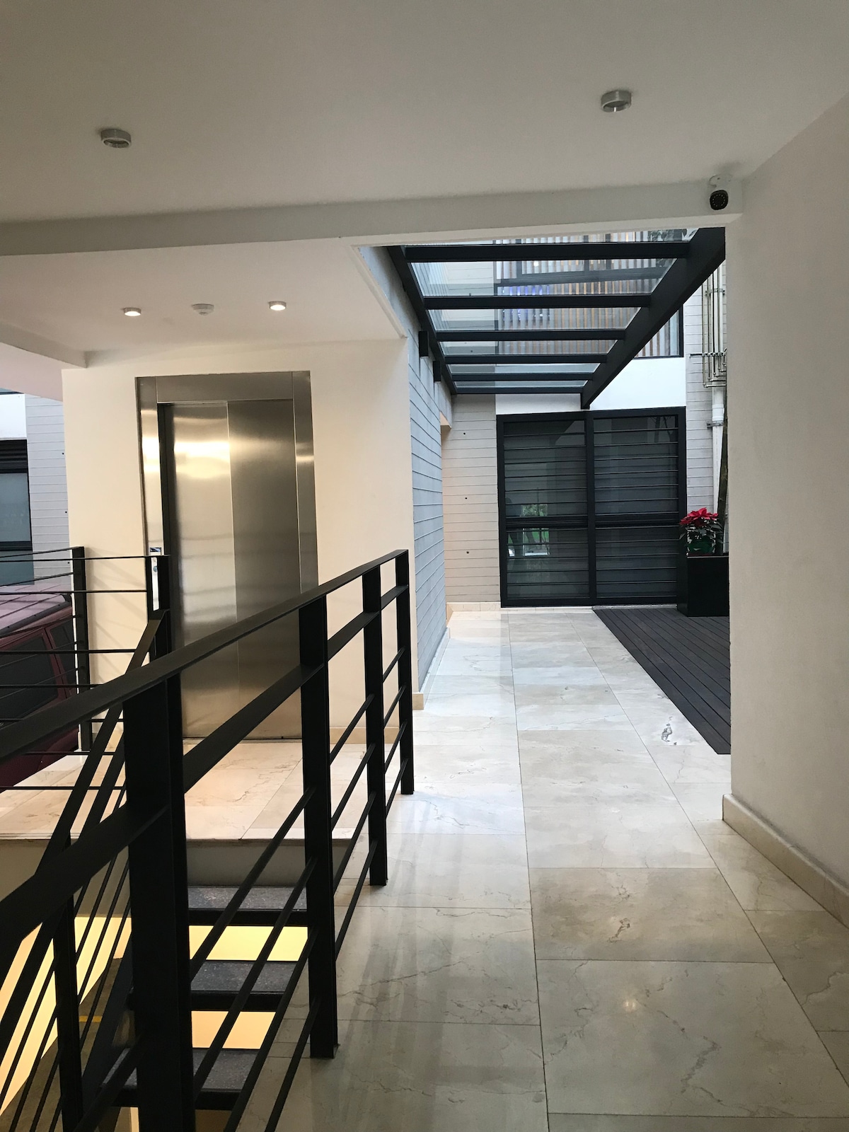 A well-lit hallway features a sleek elevator and modern railing. Natural light floods the space through large windows, illuminating the smooth marble flooring. Potted plants add a touch of greenery, while a sliding glass door leads to an outdoor area, enhancing the inviting atmosphere.