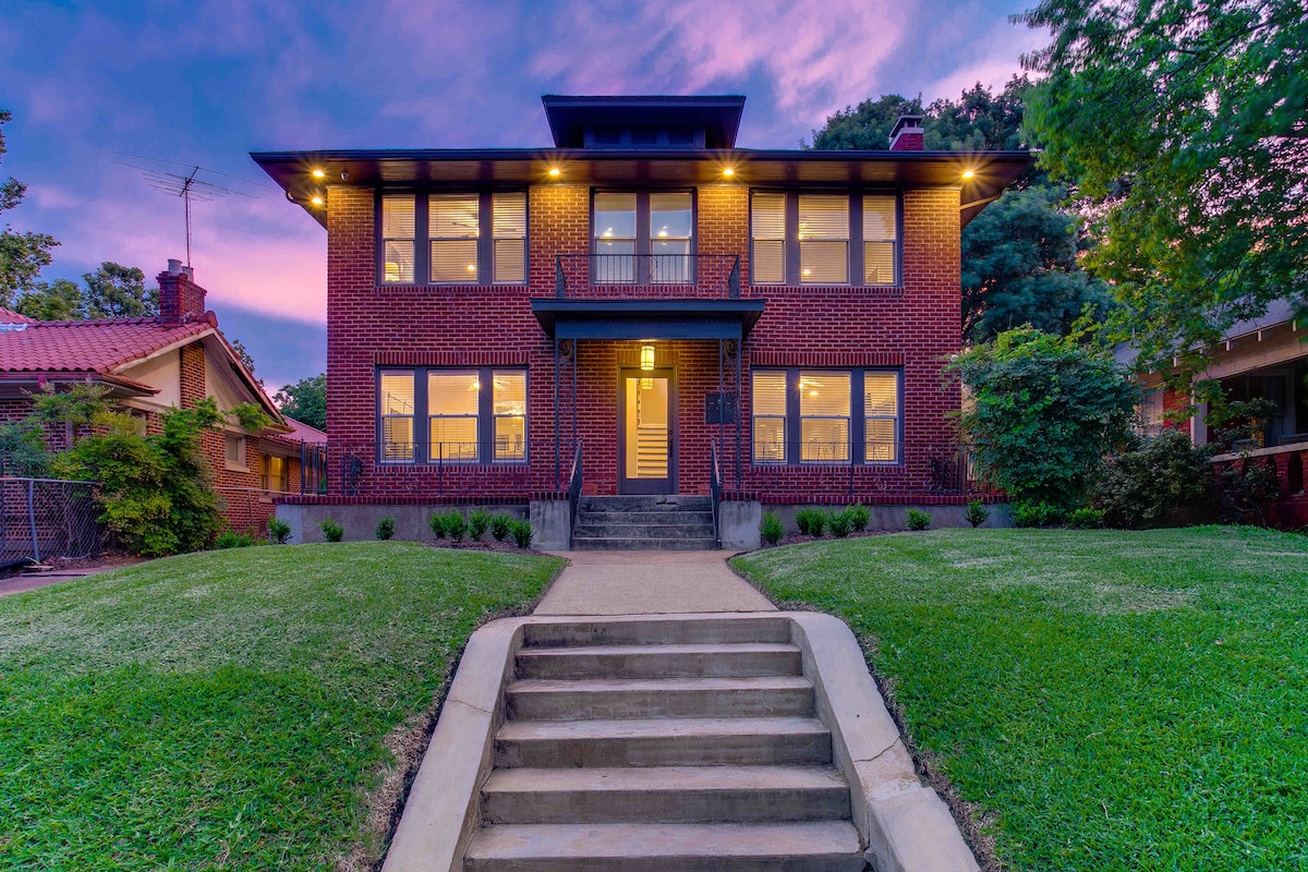 The historic brick exterior of the home is showcased against a colorful sunset, highlighting the large windows and inviting entrance. A flight of stairs leads to the front door, flanked by well-maintained greenery and soft outdoor lighting.