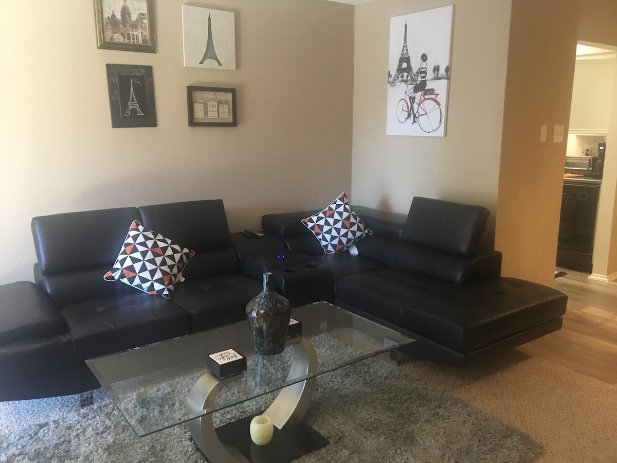 A modern living area features a black leather sectional sofa adorned with colorful accent pillows. A sleek glass coffee table sits on a soft rug, creating a comfortable gathering space. Wall art depicting iconic French landmarks enhances the ambiance, while a glimpse of the kitchen is visible in the background.