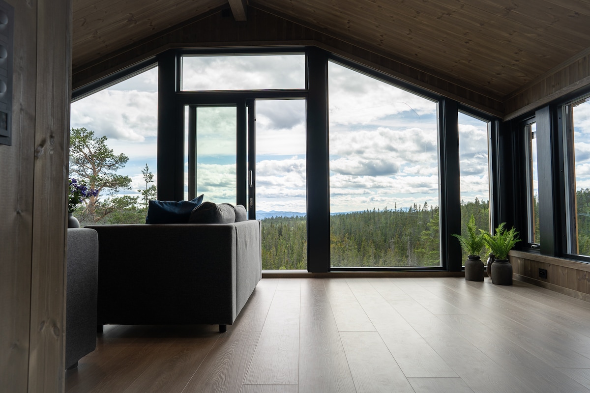 Large windows frame a panoramic view of the surrounding forest, allowing natural light to fill the space. A modern gray sofa is positioned to face the scenic landscape, complemented by small potted plants on the floor in the foreground.