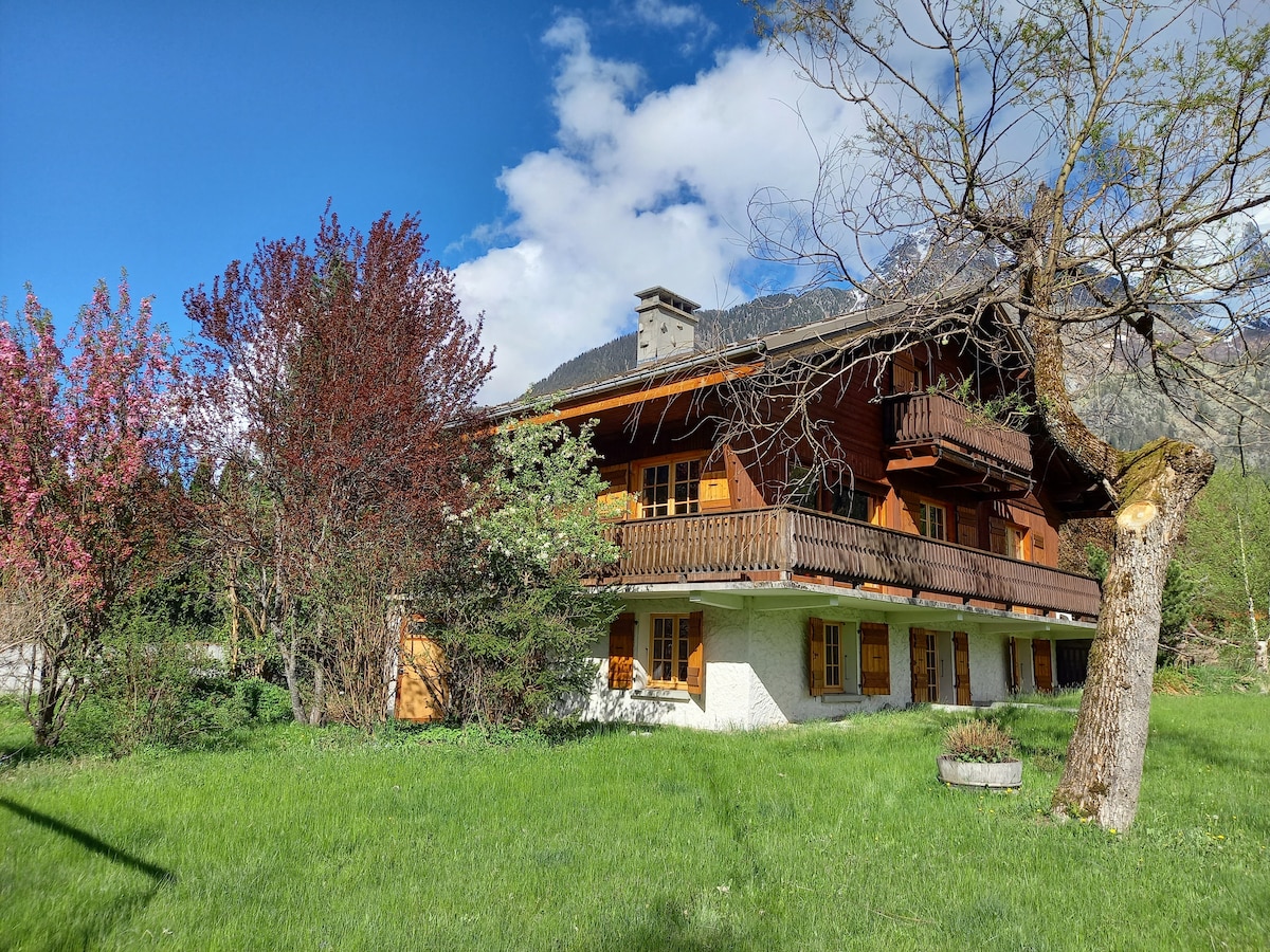 The rustic chalet is surrounded by lush green grass and vibrant flowering trees. A wooden balcony extends from the upper level, while large windows provide an inviting look. The mountainous backdrop is visible beyond the structure, contributing to the serene setting.